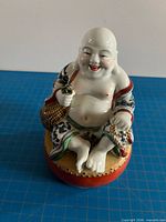 Front view of porcelain Laughing Buddha figurine seated on orange base