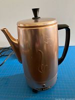 Front view of copper-tone electric percolator showing handle, spout, lid knob and embossed motif