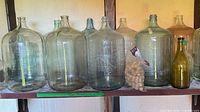 Shelf holding multiple large clear and tinted glass carboys, bag of corks and small amber jug visible