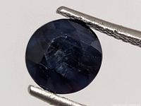 Round deep blue faceted sapphire held in tweezers, angled view