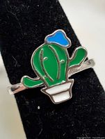 Front view of silver-tone ring showing green cactus, blue flower, white pot enamel design on black display stand
