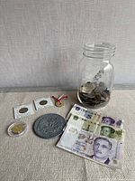 Jar of mixed coins, two Spanish coins in flips, novelty coin, gold-tone capped coin, medal, stack of banknotes