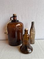 Front view of all three bottles: large amber jug with rope and cork, amber Liberty Bell bottle with cork, Coca-Cola contour bottle