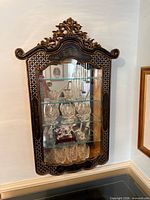 Front view of cabinet showing carved crest, gold accents, glass door, glass shelves and mirrored back