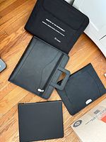 Four black office organizers laid out: faux-leather padfolio, nylon printed portfolio, neoprene sleeve, hard-cover folder