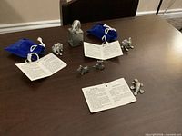 Six pewter figurines arranged on table with two blue velvet pouches and tags visible