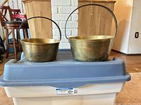 Two brass pots of different sizes with iron handles displayed on storage bin lid