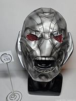 Front view of silver rubber Ultron mask with red mesh eyes on stand