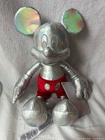 Front view of iridescent silver Mickey Mouse plush with red shorts and tag