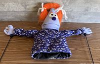 Front view of blue foam head puppet with orange yarn hair, purple swirl shirt, arm rods visible