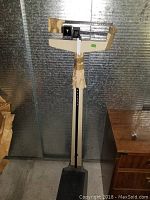Full view of the vintage doctor scale, showing the base platform, vertical column, and balance beam wrapped in protective paper and tape.