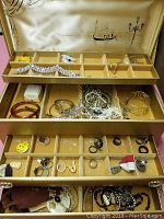 Wide shot of the jewelry box open showing three levels of compartments with assorted jewelry inside including bracelets, rings, necklaces, and pins.