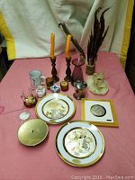 Vintage lot showing various decorative items including porcelain plates, framed artwork, brass dish, small jars, and candlesticks on a pink cloth background.