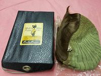 Photo of the black textured case showing a label with a Pussycat cat illustration and the brand Andy Carver next to the wig hat with light brown hair and green mesh lining.