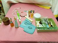 Overview of the vintage health and beauty lot showing various items on a pink surface including perfume bottles, blue cloth pads, a green organizing tray, and a battery-operated probe device.