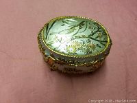 Closed oval music box showing bird and floral design on lid with gold-tone decorative accents on sides.