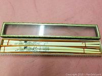 Set of four white chopsticks with black detailed illustrations in ornate greenish-gold rectangular box with clear top