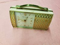 Front view of small rectangular Phinney Walker alarm clock, white clock face with gold numbers and lattice pattern accents, gold-tone handle on top.