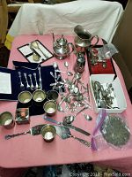 Photo showing a variety of vintage silver or silver-plated utensils, serving pieces, and small cups on a pink tablecloth.