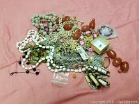 Photo shows a mix of jewelry parts including multiple strands of beads in white, green, pink, and brown hues, various silver-tone chains, silver-tone pendants, and loose beads. Some items are still connected, others are loose and tangled.