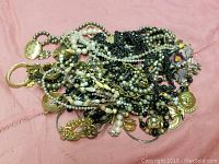 Tangled assortment of various necklaces and bracelets with metal wear parts and beads on a pink fabric background.