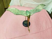 Full front and top angle showing metal bird with wings spread wide on a pink cloth background, mounted on a round black base.