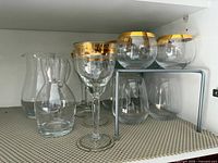 Shelf with clear carafes, one gold-rim stemmed goblet, gold-rim stemless glasses and clear stemless tumblers