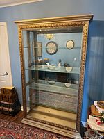 Front view of gold curio cabinet showing carved frame, glass door, shelves, mirrored back