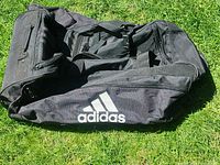 Full view of black Adidas wheeled duffel lying on grass, showing logo and overall condition