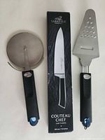 Full view of three kitchen tools: boxed Sabatier chef’s knife, pizza cutter, perforated server