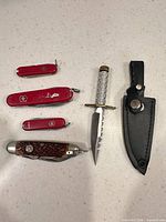 Group of five knives: three red Swiss Army pocket knives, brown jigged bone folder, silver-handled fixed blade with sheath