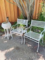 Two white metal mesh chairs, wicker planter stand, small glass light visible