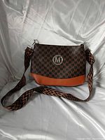 Front view of handbag showing monogram pattern, logo medallion, orange-tan base, and woven strap