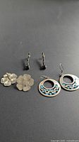 Three pairs of sterling earrings laid out: flower clip-ons, black stone drops, blue inlay rounds