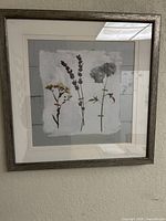 First framed pressed-flower art showing three botanical stems, silver frame and cream mat visible