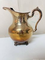 Front three-quarter view of ornate silver-plated water pitcher showing handle, spout, feet and tarnish
