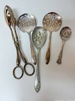 Five vintage silver plated utensils: two pierced serving spoons, embossed fruit spoon, small condiment spoon, sugar tongs