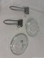 Two clear glass lids with knob handles and two detachable metal saucepan handles on white surface
