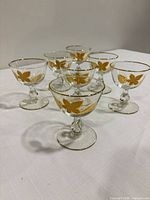 Seven clear glass champagne coupes arranged together showing gold leaf trim and stems