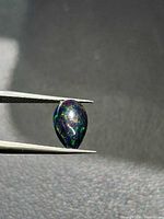 Pear cabochon opal held in tweezers, showing dark body and green, blue flashes