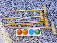 Six wooden mallets, four colored balls, and bundled white wickets laid on gravel
