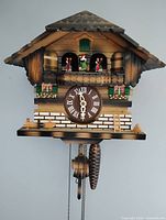 Front view of wooden chalet cuckoo clock showing dial, balcony dancers and pinecone weight