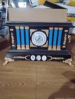 Front view of black wood mantel clock with blue panels, scenic dial, gold accents and feet