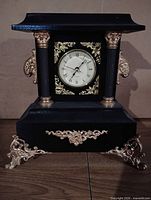 Front view of black and gold handmade mantel clock showing dial, columns and decorative scroll feet