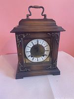 front view of wooden mantel clock showing dial, glass door, brass-tone appliqués, top handle