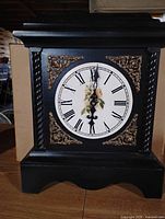 Front view of black wooden mantel clock with Roman numeral dial, floral center, gold corner ornaments, rope-style columns