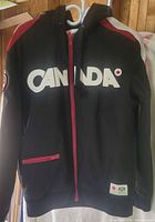 Front view of black Canada Olympic full-zip hoodie showing CANADA lettering, red zipper, front zip pocket