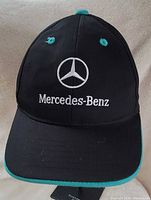Front view showing embroidered Mercedes-Benz logo and text on black cap with teal trim