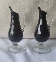 Front view showing both black amethyst glass pitchers with clear handles and pedestal bases