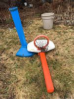Disassembled youth basketball hoop showing blue base and pole, white backboard, orange rim, black net and orange pole segment on lawn